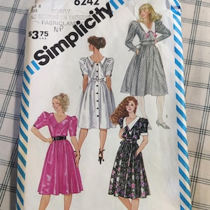 May include: Simplicity sewing pattern number 6242 for a dress with puffed sleeves. The pattern shows three different styles of dresses: a white dress with a button-up front, a pink dress with a black belt, and a floral print dress with a white collar.