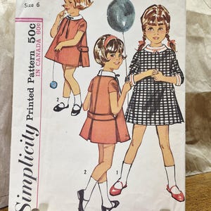 Op de afbeelding: Een vintage Simplicity naaipatroon-envelop, nummer 5809, voor een kinderjurk in maat 6. De omslag toont illustraties van drie kinderen in jurken met korte mouwen en witte kragen, waaronder één in een zwart-wit ruitjespatroon.