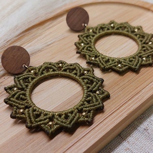 May include: Olive green macrame earrings with a circular design and small gold beads. The earrings have a wooden stud backing. The earrings are displayed on a wooden tray.