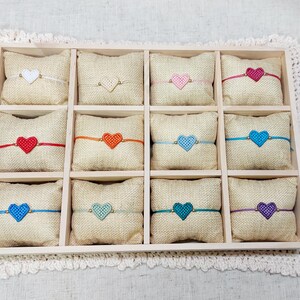 May include: A wooden tray with twelve small, square burlap pillows, each adorned with a heart-shaped embellishment. The hearts are in various colors, including red, pink, blue, and purple. The tray sits on a white, lace-edged surface.