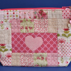 May include: A pink and white patchwork zippered pouch with a heart-shaped appliqué. The pouch has a zipper closure and a pink charm with hearts.