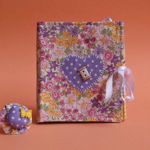 May include: A purple and white floral fabric covered journal with a white button and a purple heart on the front. The journal has a white ribbon tied around it.