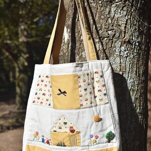 Może przedstawiać: Biała torba typu tote z płótna z panelami z tkaniny w żółto-białe wzory. Torba ma wzór domu z czerwonym ptakiem i dwiema małymi postaciami z przodu. W dolnych rogach torby znajdują się żółte kokardki.