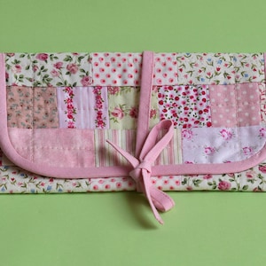 Może przedstawiać: Różowa torebka z tkaniny w patchworku z różową wstążką zawiązywaną. Torebka jest wykonana z różnych tkanin kwiatowych i wzorzystych w odcieniach różu i bieli.
