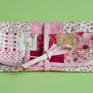 Może przedstawiać: Torebka z patchworku z różową i białą wstążką w kropki zawiązaną wokół niej. Torebka jest wykonana z różnych tkanin kwiatowych i w kropki w odcieniach różowego, czerwonego i białego.