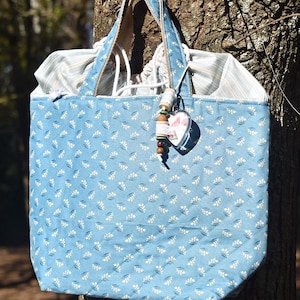 May include: A light blue tote bag with a white floral pattern. The bag has a brown handle and a heart-shaped charm with beads.