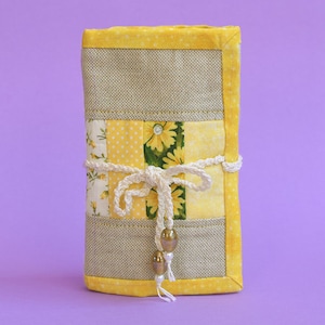 May include: A yellow and beige fabric pouch with a white crocheted tie and gold beads. The pouch is made of patchwork fabric with floral and polka dot patterns.
