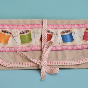 Op de afbeelding: Een beige stoffen rolhoes met roze stippenbies en een roze lint. De hoes heeft een print van kleurrijke spoelen garen met naalden.