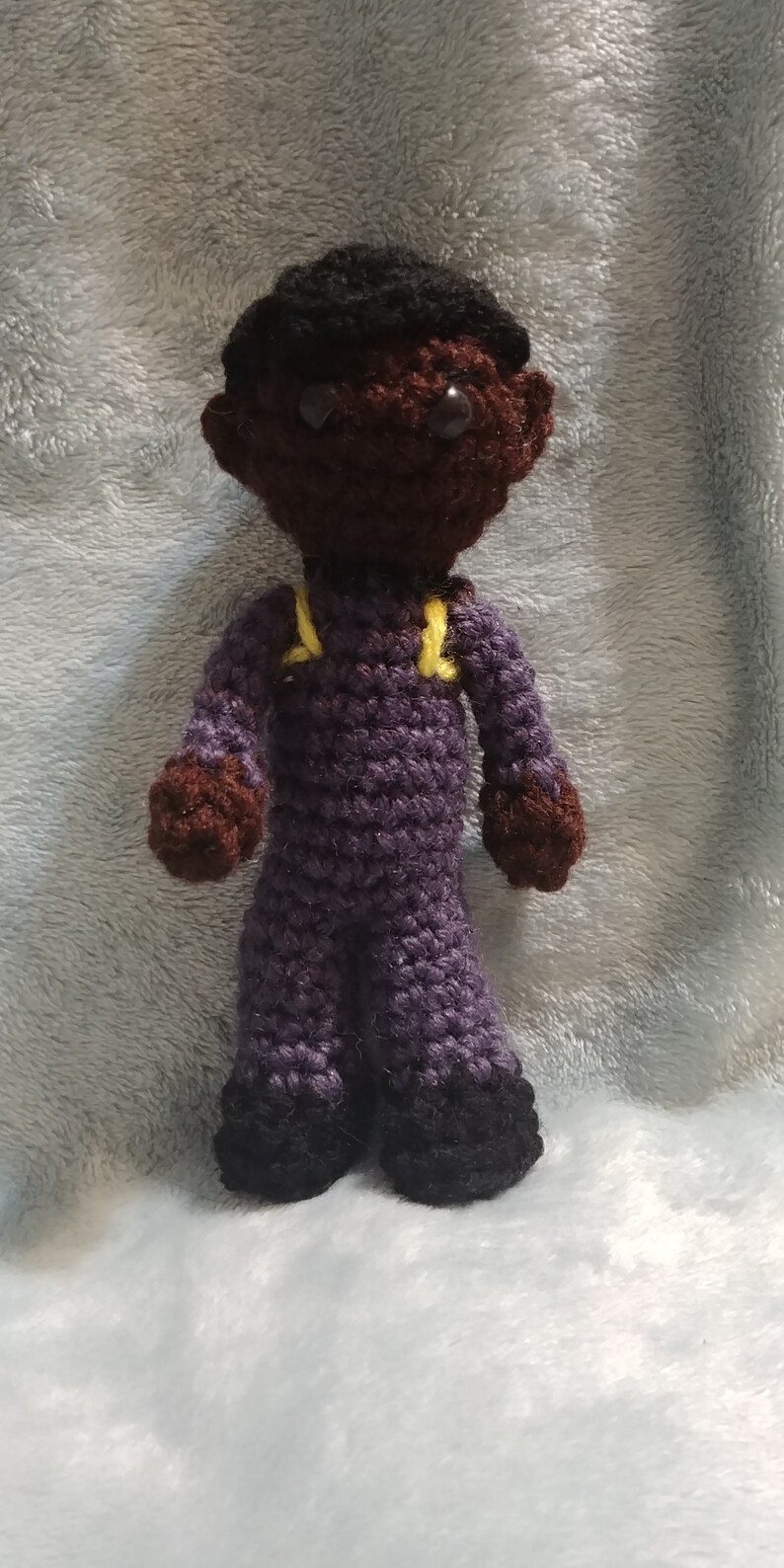 Pode incluir: Uma boneca de croch&ecirc; com pele marrom, cabelo preto e macac&atilde;o roxo. A boneca tem al&ccedil;as amarelas no macac&atilde;o e sapatos pretos.