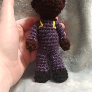 Pode incluir: Uma boneca de croch&ecirc; com pele marrom, cabelo preto e roupa roxa. A boneca est&aacute; usando um uniforme amarelo e preto com sapatos pretos.