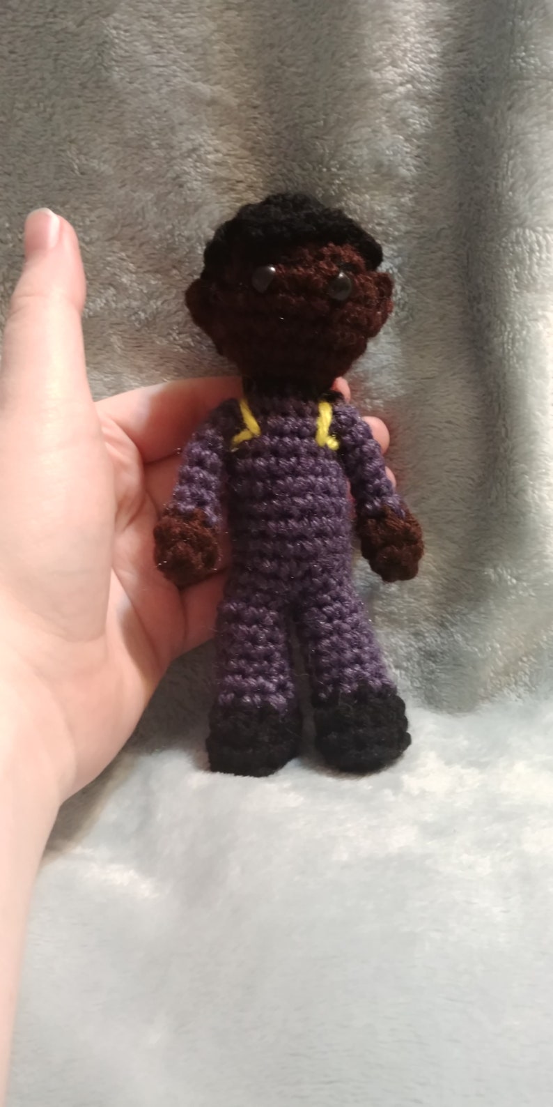 Pode incluir: Uma boneca de croch&ecirc; com pele marrom, cabelo preto e roupa roxa. A boneca est&aacute; usando um uniforme amarelo e preto com sapatos pretos.