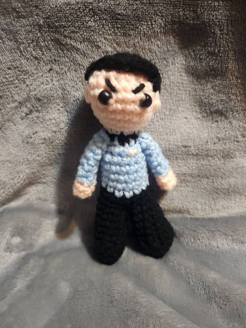 K&ouml;nnte beinhalten: Eine geh&auml;kelte Puppe mit schwarzen Haaren, einem blauen Hemd und schwarzen Hosen. Die Puppe hat einen ernsten Ausdruck mit zusammengezogenen Augenbrauen und schwarzen Augen.