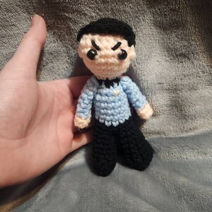 K&ouml;nnte beinhalten: Eine geh&auml;kelte Figur einer Person mit schwarzen Haaren, einem blauen Hemd und einer schwarzen Hose. Die Figur hat einen strengen Ausdruck mit zusammengezogenen Augenbrauen und einem Stirnrunzeln.