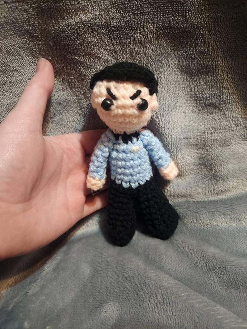 K&ouml;nnte beinhalten: Eine geh&auml;kelte Figur einer Person mit schwarzen Haaren, einem blauen Hemd und einer schwarzen Hose. Die Figur hat einen strengen Ausdruck mit zusammengezogenen Augenbrauen und einem Stirnrunzeln.