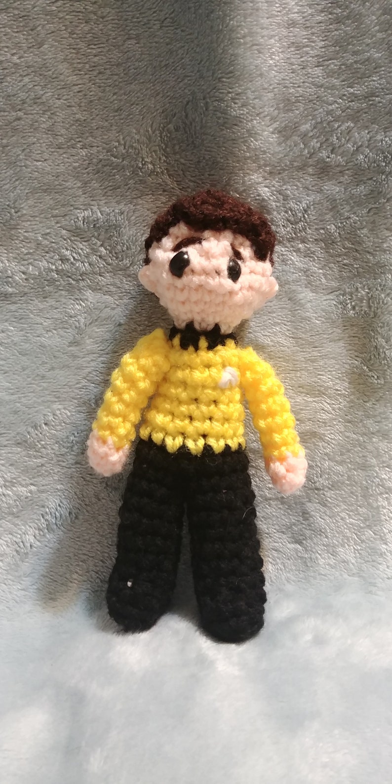 May include: A crocheted doll wearing a yellow shirt with a black collar and a black star, black pants, and brown hair.