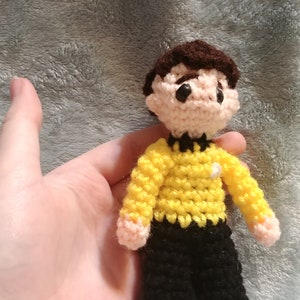 May include: A crocheted doll of a person wearing a yellow shirt with a black collar and black pants. The doll has brown hair and black eyes.