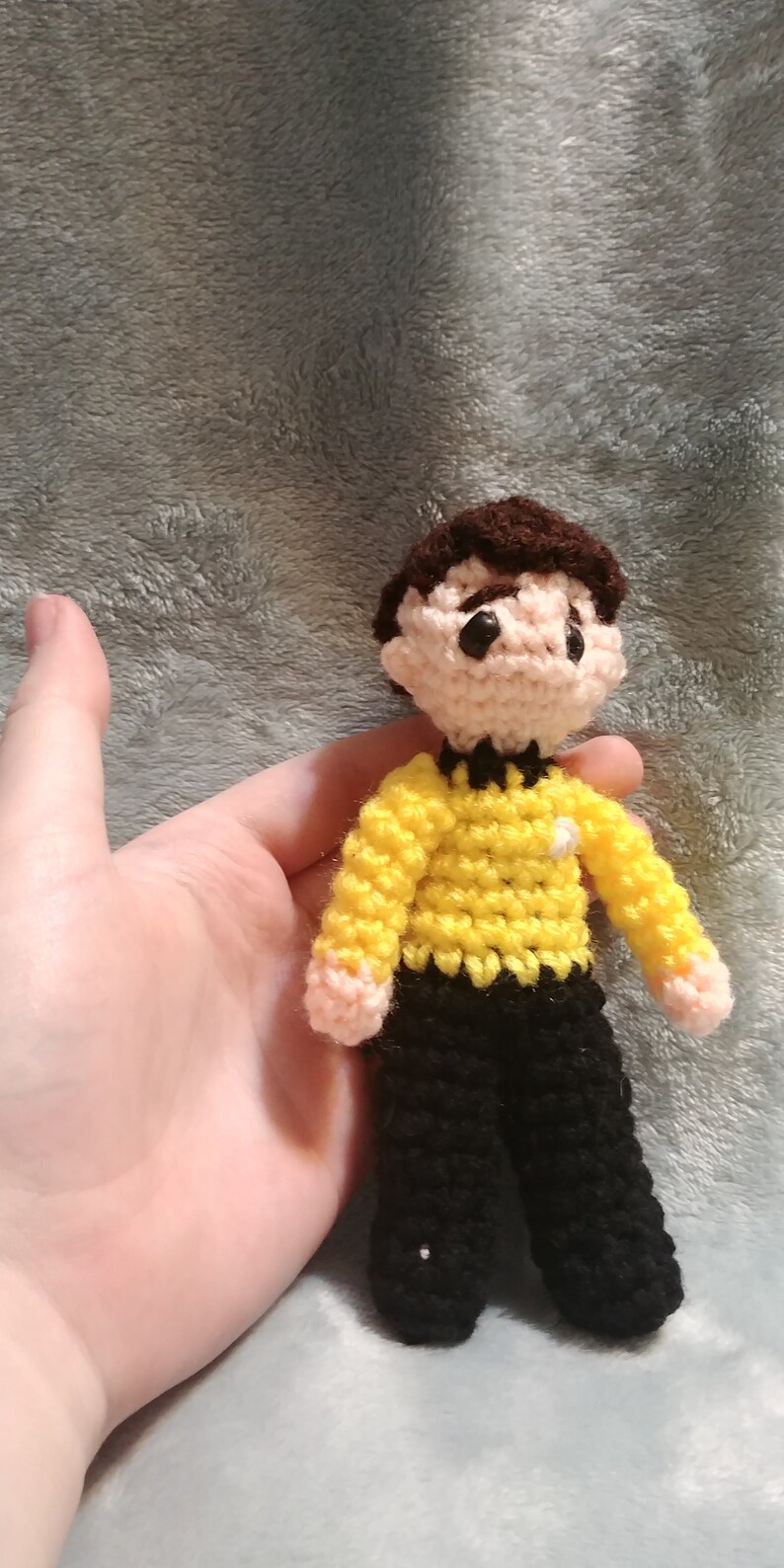 May include: A crocheted doll of a person wearing a yellow shirt with a black collar and black pants. The doll has brown hair and black eyes.