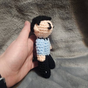 K&ouml;nnte beinhalten: Eine geh&auml;kelte Puppe mit schwarzen Haaren, einem hellblauen Hemd und schwarzen Hosen. Die Puppe steht auf einer grauen, flauschigen Oberfl&auml;che.