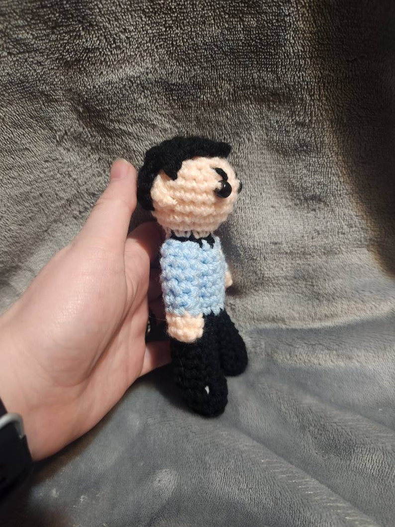 K&ouml;nnte beinhalten: Eine geh&auml;kelte Puppe mit schwarzen Haaren, einem hellblauen Hemd und schwarzen Hosen. Die Puppe steht auf einer grauen, flauschigen Oberfl&auml;che.