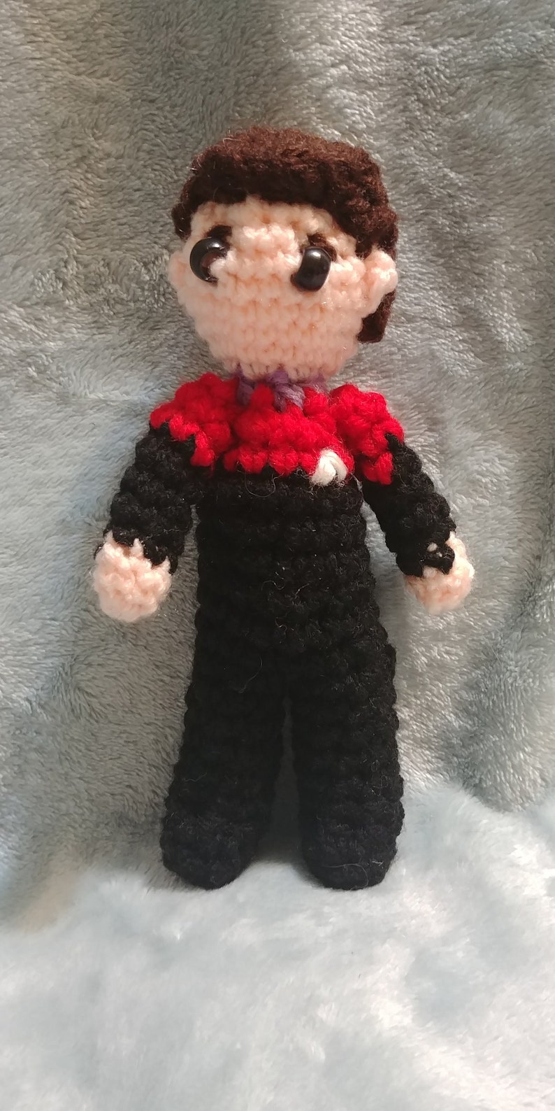 May include: Crocheted amigurumi doll of a person wearing a black uniform with a red and white shirt. The doll has brown hair and black eyes.