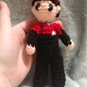 May include: Crocheted figure of a person wearing a black uniform with red and purple accents. The figure has brown hair and a neutral facial expression.