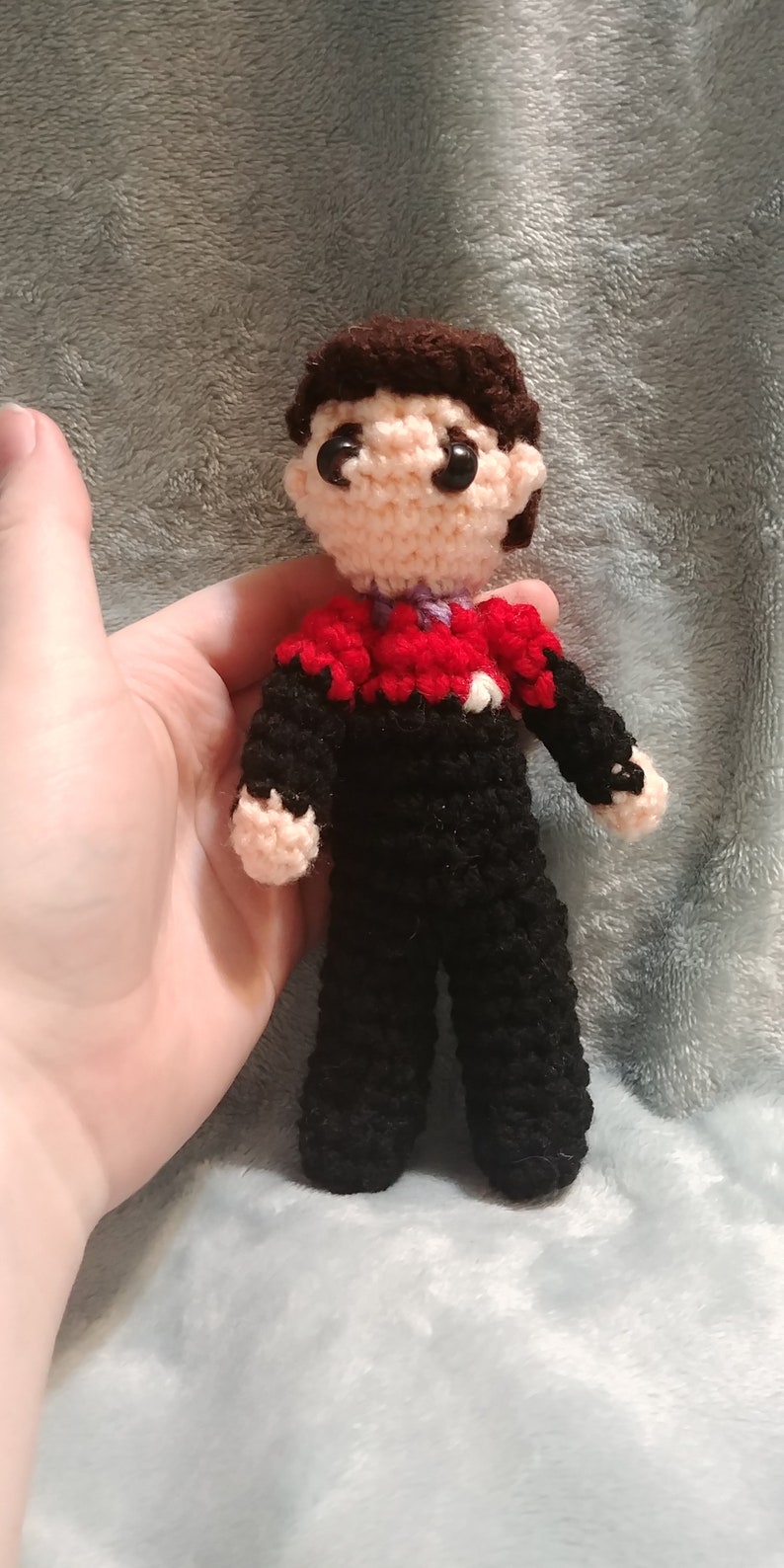 May include: Crocheted figure of a person wearing a black uniform with red and purple accents. The figure has brown hair and a neutral facial expression.
