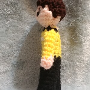 May include: Crocheted figure of a person wearing a yellow shirt and black pants. The figure has brown hair and a pink face.
