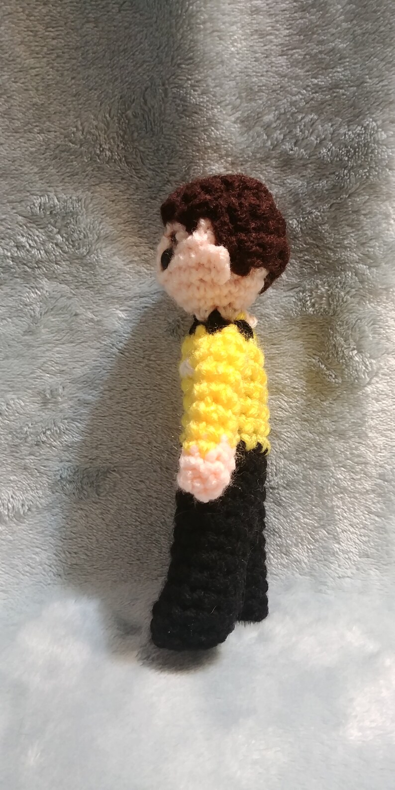 May include: Crocheted figure of a person wearing a yellow shirt and black pants. The figure has brown hair and a pink face.