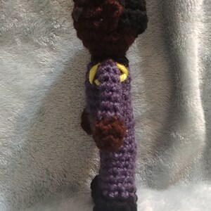 Pode incluir: Boneca de croch&ecirc; com pele marrom, cabelo preto e roupa roxa com detalhes amarelos. A boneca est&aacute; em p&eacute; sobre uma superf&iacute;cie cinza e felpuda.