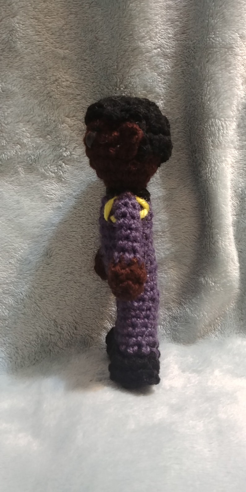 Pode incluir: Boneca de croch&ecirc; com pele marrom, cabelo preto e roupa roxa com detalhes amarelos. A boneca est&aacute; em p&eacute; sobre uma superf&iacute;cie cinza e felpuda.