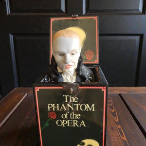 May include: A vintage Jack-in-the-Box toy featuring the Phantom of the Opera. The black box has the title in white and red lettering, with a red rose and a mask graphic. The figure has a white mask and a black coat.