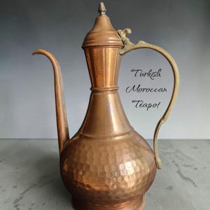 May include: A copper-colored teapot with a long spout and a curved handle. The body of the teapot has a textured, hammered design. The handle and the top finial are gold-colored. The text "Turkish Moroccan Teapot" is visible.