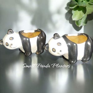 May include: Two ceramic panda planters with black and white coloring. The planters have a small opening for plants and are shaped like pandas. The text "Small Panda Planters" is written in cursive below the planters.