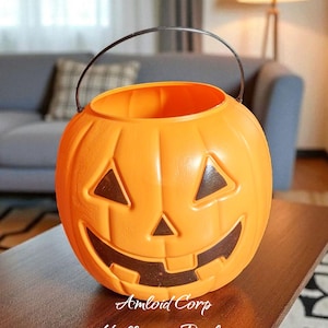 May include: Orange plastic Halloween bucket with a carved jack-o'-lantern face. The bucket has a black metal handle and the text "Amlord Corp Halloween Bucket" on the front. The bucket is sitting on a brown table.
