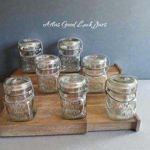 May include: A collection of clear glass jars with metal handles and lids, arranged on a wooden tray. The jars have a square shape with a floral design and the text "Atlas Good Luck Jars". The background is a solid gray color.