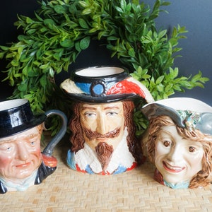 May include: Three ceramic mugs with painted faces. The mugs are in the shape of a man with a black hat, a man with a red and blue hat, and a woman with a blue and white hat. The mugs are on a woven surface.