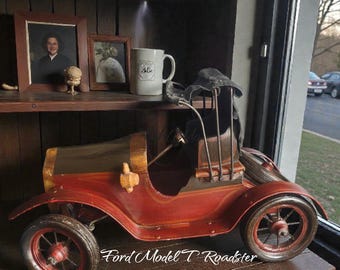 May include: A vintage-style, red and brown Ford Model T Roadster model sits on a wooden shelf. The model car features detailed wheels, a black top, and gold accents. The shelf also displays framed photos and a mug.