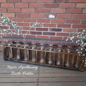 May include: A rustic wooden bottle holder with ten clear glass bottles. The holder is made of stained wood and sits on a wooden surface. The text "Rustic Apothecary Bottle Holder" is visible. The background is a brick wall.