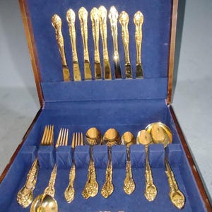 May include: A gold-toned stainless steel flatware set from Japan, presented in a wooden box with a blue velvet interior. The set includes knives, forks, and spoons with ornate handles. The box lid features the text Stainless Japan Flatware Set.