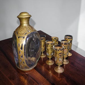May include: A vintage glass decanter with a gold-colored metal design and matching shot glasses. The decanter has a rounded shape with a stopper. The shot glasses have a similar gold design and are on small pedestals. The set is displayed on a dark wooden surface.