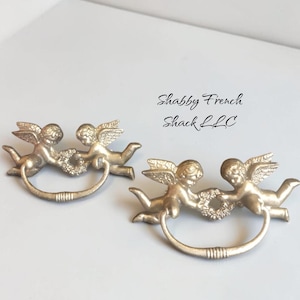 May include: Two metallic gold-toned drawer pulls, each featuring a pair of winged cherubs holding a central floral wreath. A curved handle hangs from the cherubs. The text "Shabby French Shack LLC" is visible in the upper right corner of the image.