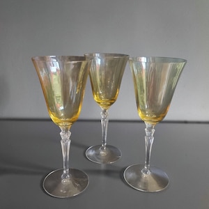 May include: Three matching wine glasses with translucent amber-colored bowls and clear, faceted glass stems. Each glass features a wide, flared rim, a tapered body, and a circular base. The stems have a decorative, multi-sided design.