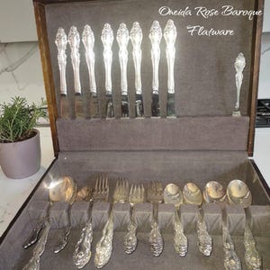 May include: A set of Oneida Rose Baroque flatware stored in a dark wooden case with a grey velvet interior. The collection includes silver-toned knives, forks, and spoons with ornate floral handles, arranged in dedicated slots within the box.