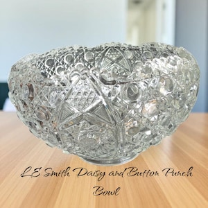 May include: A clear, cut-glass punch bowl with a detailed Daisy and Button pattern. The bowl has a wide, oval shape and sits on a wooden surface. The text "LE Smith Daisy and Button Punch Bowl" is below the bowl.