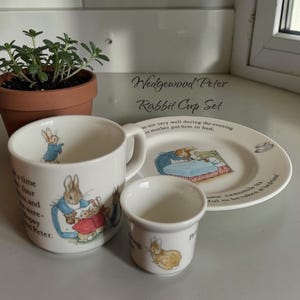 May include: A white ceramic Peter Rabbit cup set, including a mug, small cup, and saucer. The set features illustrations of Peter Rabbit and text. A small potted plant is in the background.