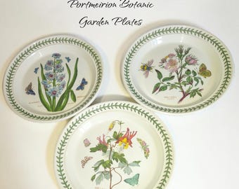 May include: Three round ceramic plates from the Portmeirion Botanic Garden collection. Each white plate features a green leaf border and a unique botanical illustration of flowers, including hyacinth, rose, and columbine, with small insects.