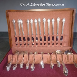 May include: A wooden storage chest containing a set of Oneida Silverplate Remembrance flatware. The set includes silver-plated knives, forks, and spoons arranged in a brown velvet-lined interior with individual slots for each piece.