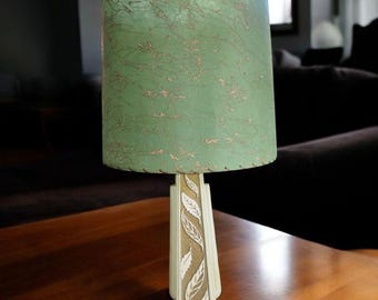 May include: Vintage table lamp with a green lampshade featuring gold-colored vein-like patterns. The lamp base is off-white with a leaf design. The lamp is placed on a wooden table, creating a warm and inviting ambiance.