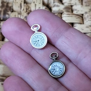 Puede incluir: Dos encantos de reloj de bolsillo en miniatura, uno plateado y otro dorado, con esferas de números romanos.