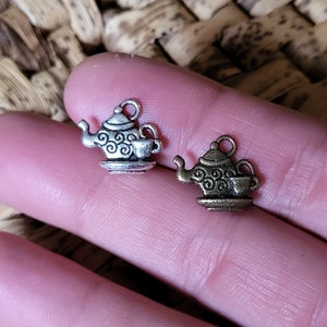 Könnte beinhalten: Zwei kleine Metallanhänger in Form von Teekannen mit passenden Tassen. Die Anhänger sind silber- und bronzefarben und weisen aufwendige Wirbelmuster auf den Teekannen auf. Die Anhänger werden in einer Hand gehalten.